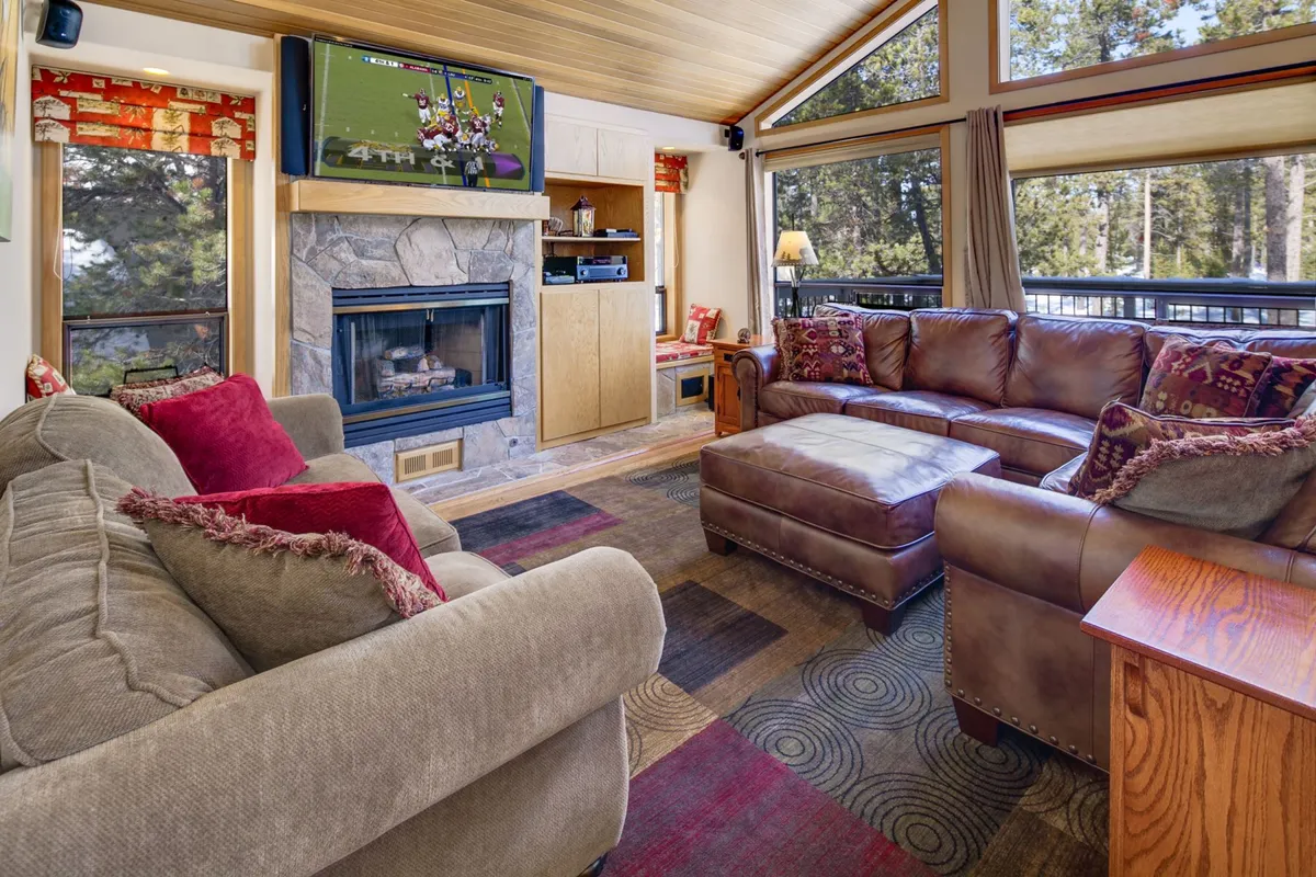 This image showcases a central living space featuring a stone-set gas fireplace, comfortable seating arranged to encourage conversation, a large television, and expansive windows that offer a captivating view of the forest outside. The hardwood floors and ample natural light create a warm and inviting atmosphere, perfectly encapsulating the gathering hub described in the section.