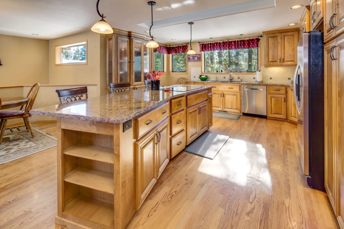 Here, the image features an open-plan kitchen and dining area. Granite countertops, custom wood cabinetry, and stainless steel appliances are visible, with a generous central island perfect for entertaining or meal prep. The adjacent dining space is surrounded by windows, further reinforcing the connection to the forest outdoors.