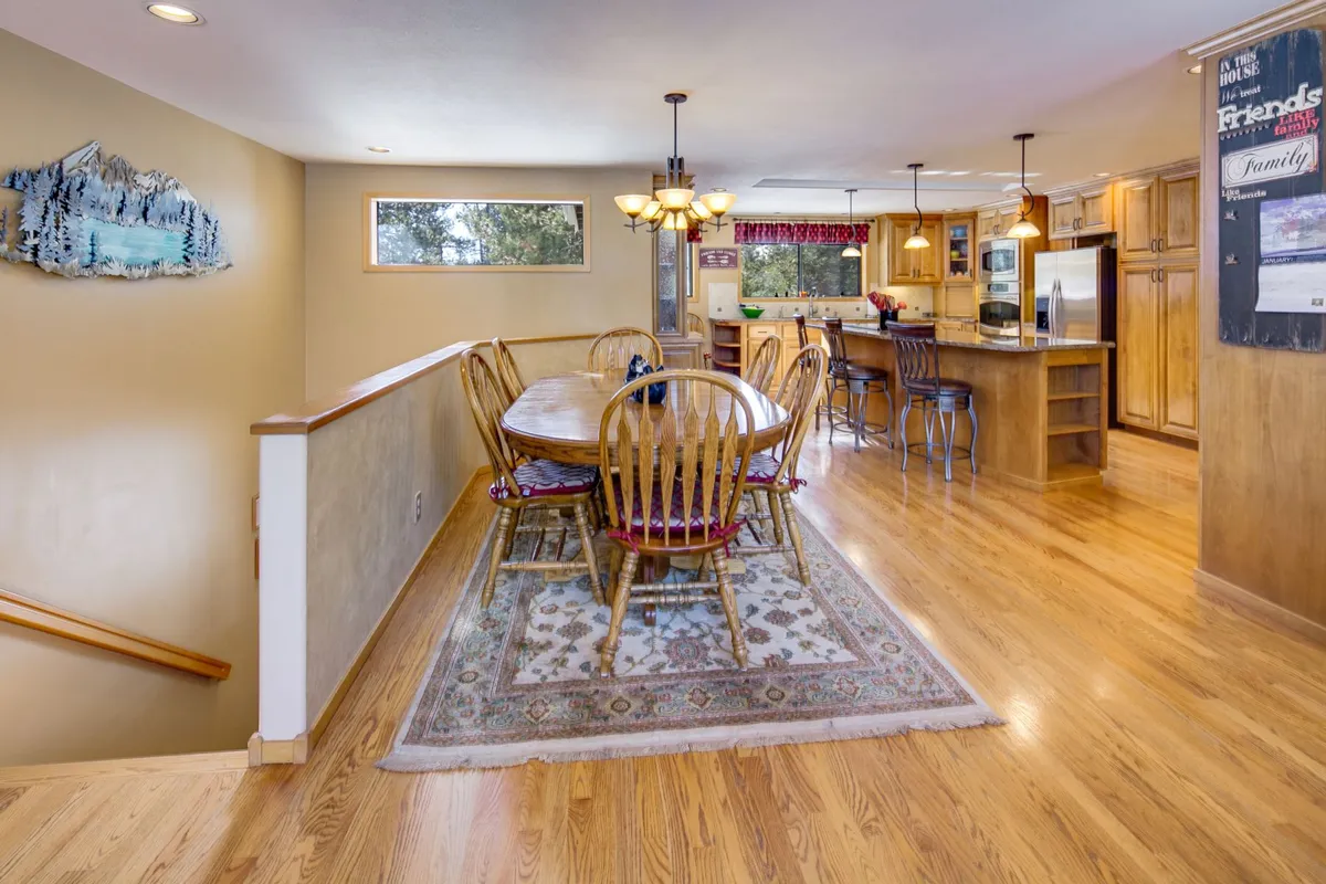 This image features a spacious dining table set in an open-concept area with a view of the kitchen, wooden railing, and decorative wall art. Natural light flows into the room, and the seating arrangement encourages group gatherings and shared meals. The visible kitchen area hints at a chef’s kitchen ideal for entertaining.