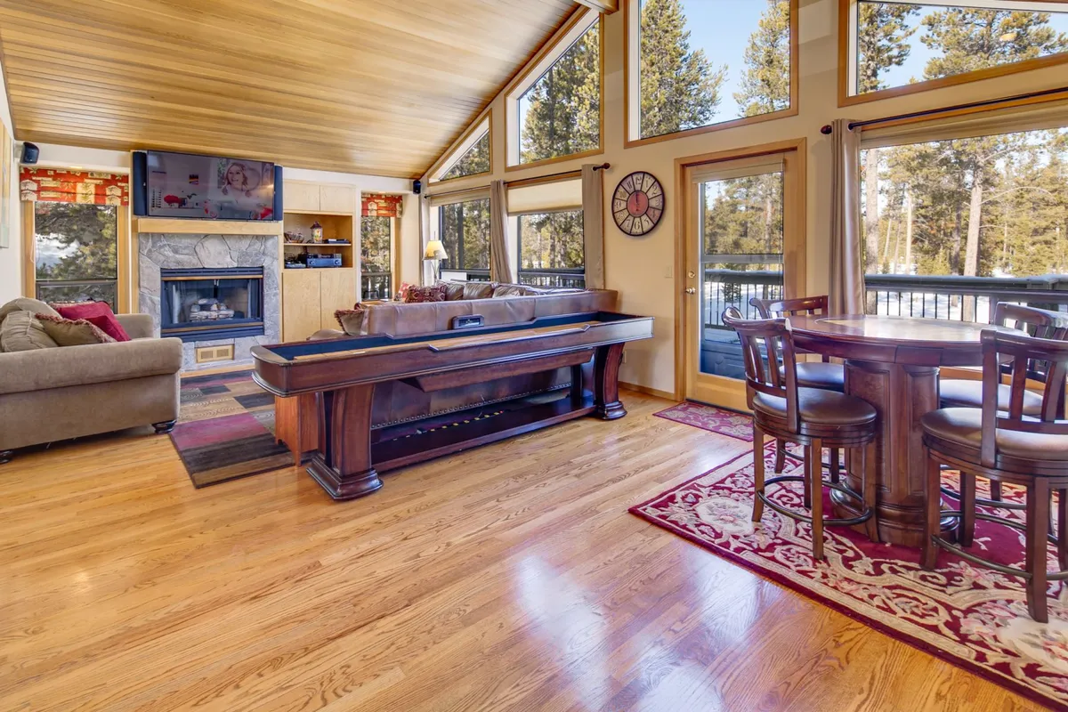 This room features a pool table as a fun centerpiece, a stone fireplace, a large dining table, and oversized windows offering glimpses of the surrounding forest. The space is designed for gatherings—be it game nights, family dinners, or relaxing by the fire.