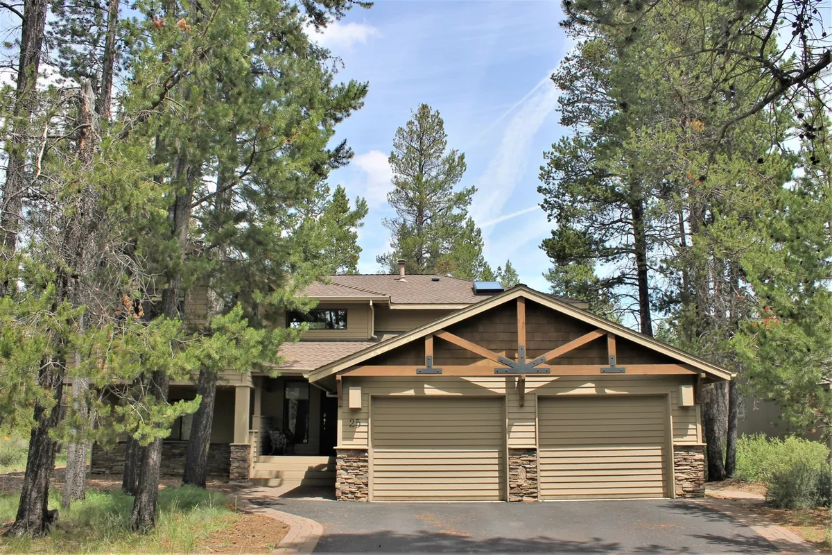 An exterior view of the home’s front entrance, featuring attractive stone accents, a spacious two-car garage, and tall trees framing the property. The inviting driveway and visible garage doors emphasize easy access for adventurers and make a welcoming first impression for guests returning from outdoor activities.