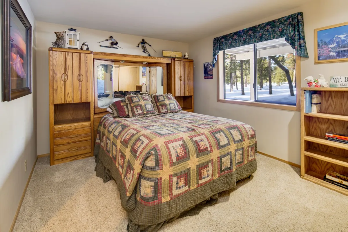 A cozy bedroom scene showing a bed with a patterned quilt, wooden furniture, and a large window offering a clear view of the outdoors. The natural light pouring in emphasizes the connection to the surrounding landscape and the serenity of private moments after active days.