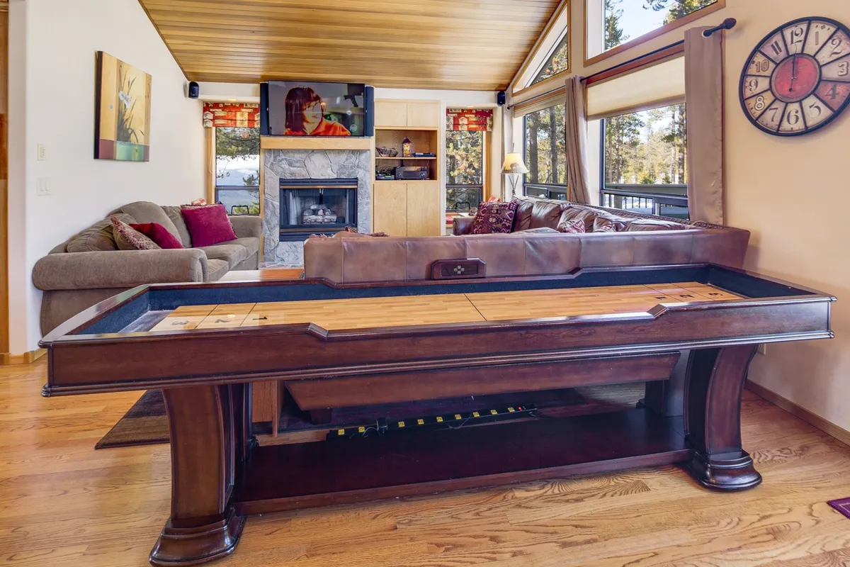 A spacious living area featuring a shuffleboard table in the foreground, a cozy sofa facing a large television, and a glowing fireplace with substantial windows letting natural light spill in. The setup invites social interaction, game nights, and movie watching, all amid a warmly furnished, inviting environment.