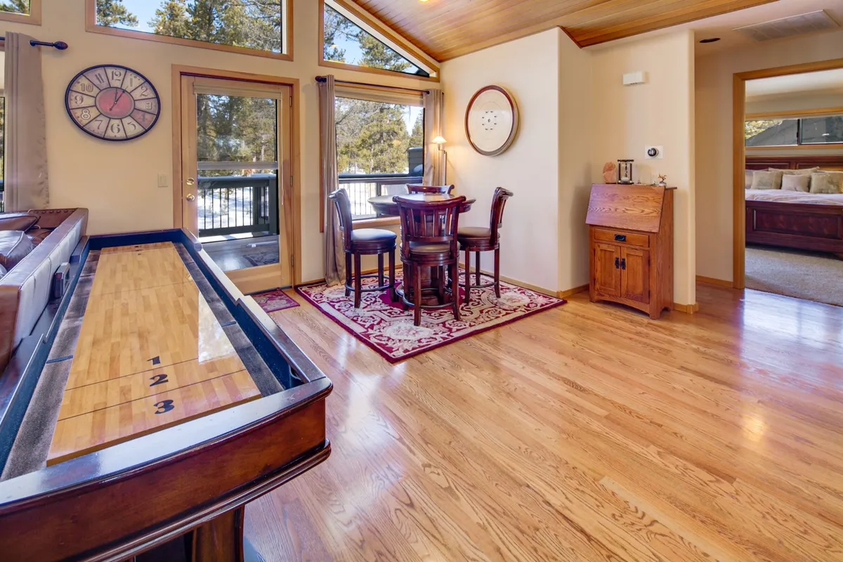 A sunlit open-plan space with a shuffleboard table, a large dining table surrounded by chairs, and oversized windows with forest views. The decor is warm, and the room feels lively and perfect for family gatherings, meals, and games.