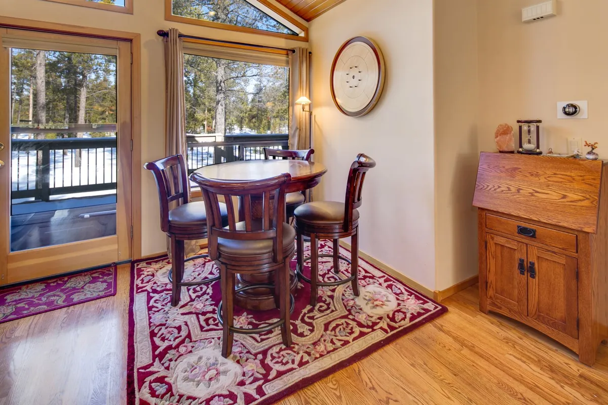 A cozy round dining table with wooden chairs sits atop a decorative rug near large windows with woodland views. The ambiance is intimate and inviting, perfect for morning coffee or evening cocktails, and the natural setting outside creates a seamless connection to the outdoors.