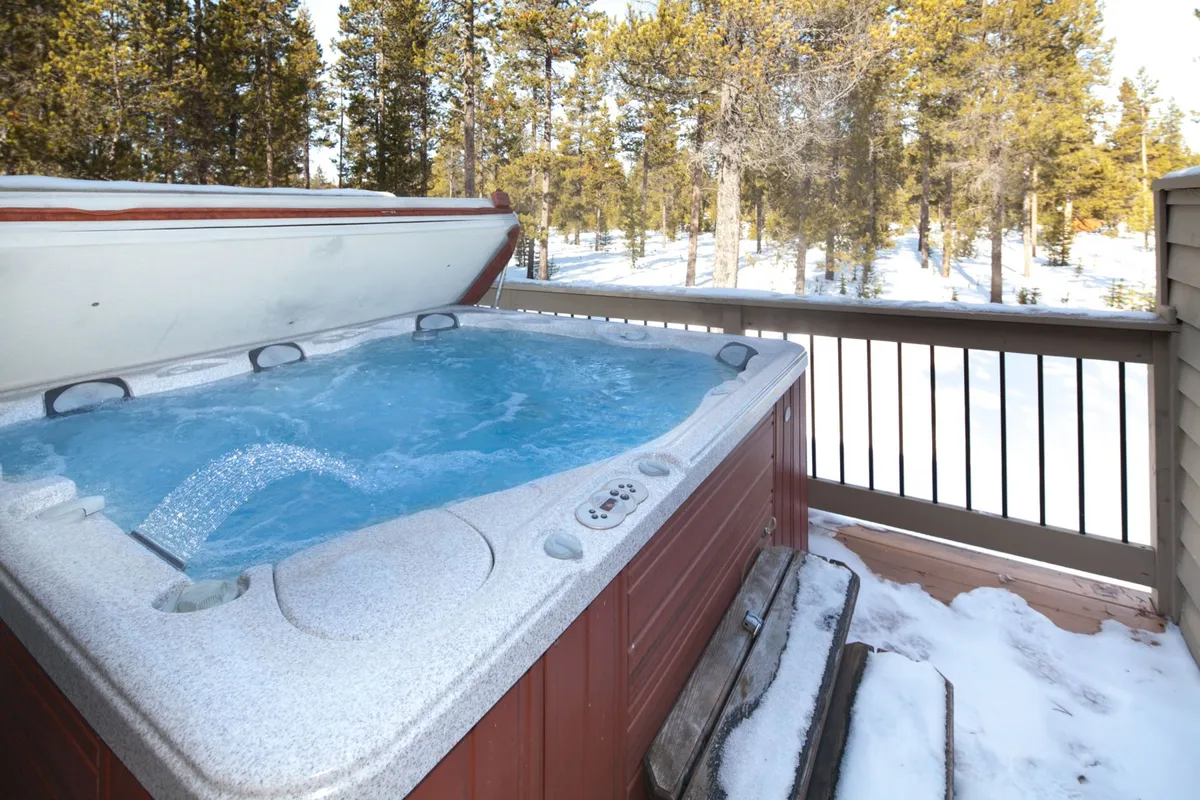This exterior image captures a private deck with a hot tub surrounded by snow-covered trees. The deck is bordered by a wooden railing, and the forest extends beyond, creating a sense of seclusion and immersion in nature. The snowy landscape highlights the tranquility and scenic beauty of the setting.